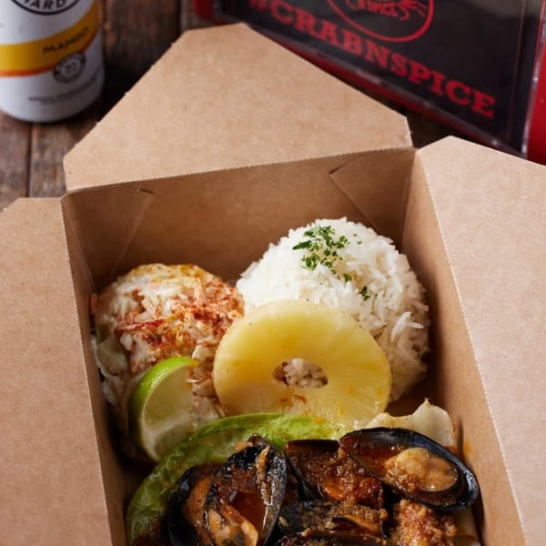 Crab N' Spice seafood meal in a takeout box with mussels, rice, coleslaw, pineapple, and lime.