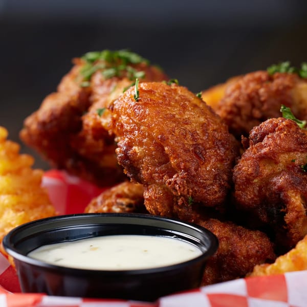 Crispy fried chicken wings with waffle fries and a side of dipping sauce in a red and white basket.