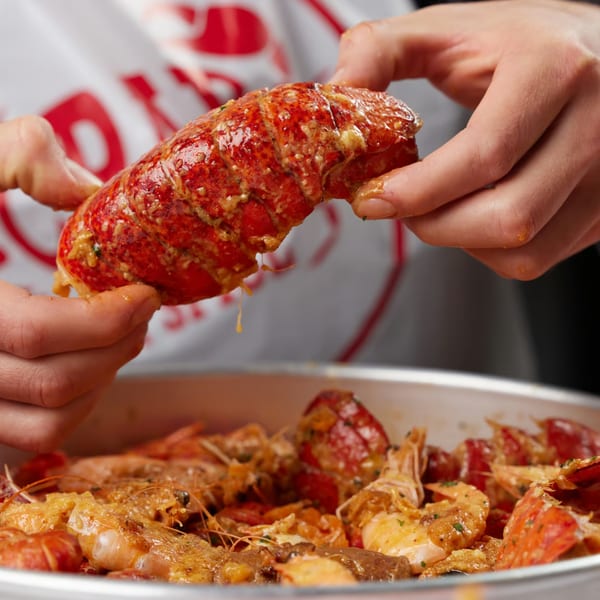 Hands holding a large, saucy lobster tail over a pan of seafood at Crab N' Spice.