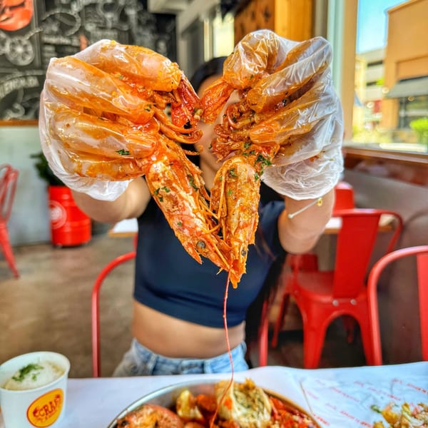 Hands in plastic gloves hold up two large, saucy prawns in a heart shape at Crab N' Spice.