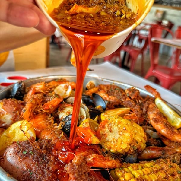 Spicy Cajun sauce pouring over a platter of Crab N' Spice seafood boil with shrimp, mussels, corn, and potatoes.
